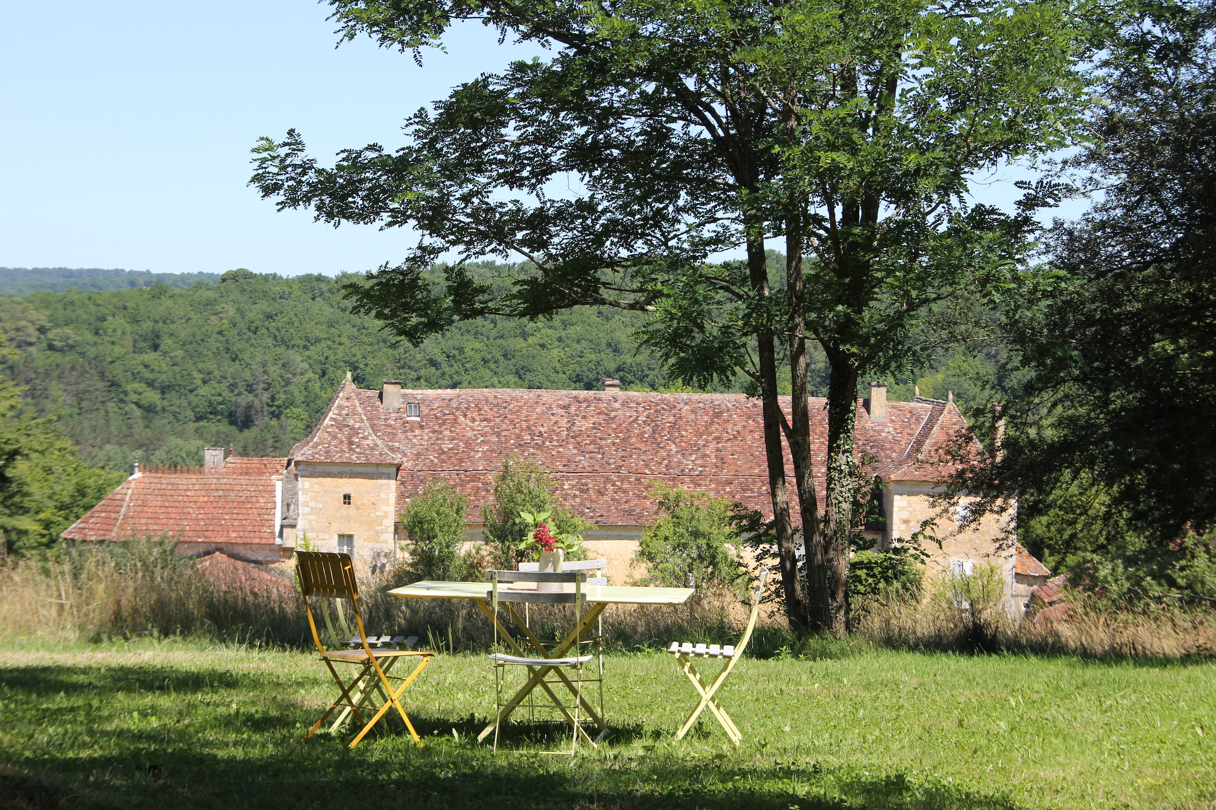 Table et chaises au premier plan dans un jardin avec un château en contrebas et arbre ombrageant une partie du jardin