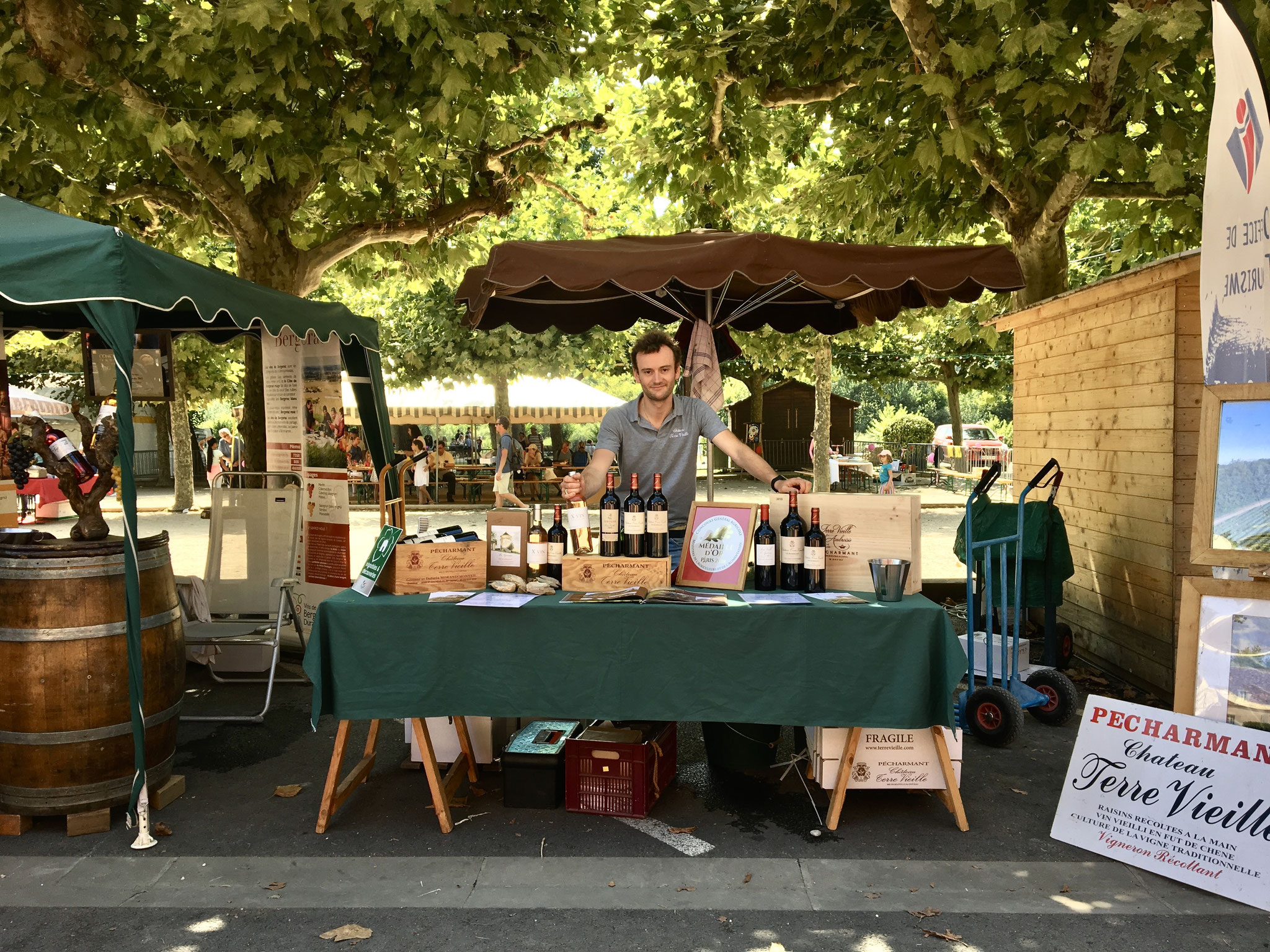 Stand de marché en extérieur avec étalage de vins du pays de Bergerac et Pécharmant. Jeune homme derrière une table présente ses vins. panneaux Château terre Vieille